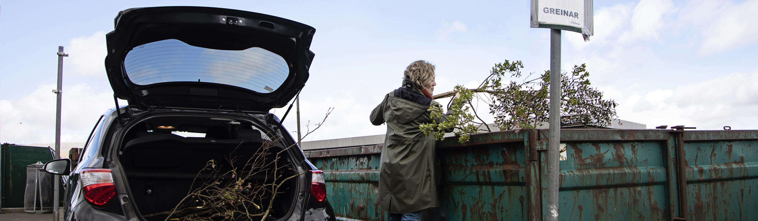A person is disposing of branches into a large green dumpster, with a car parked nearby and a cloudy sky above.