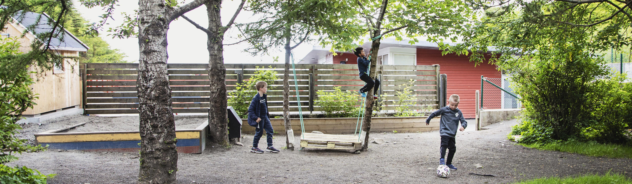 Children playing in a playground with trees, a swing, and a small soccer ball. A wooden structure is visible in the background.