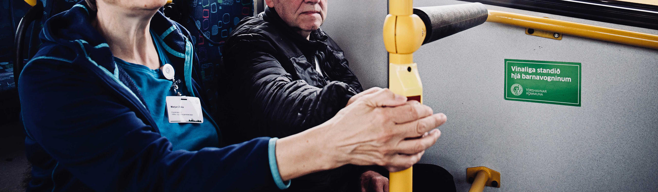 Two people sitting on a bus, one holding a yellow pole. A sign in the background is visible.