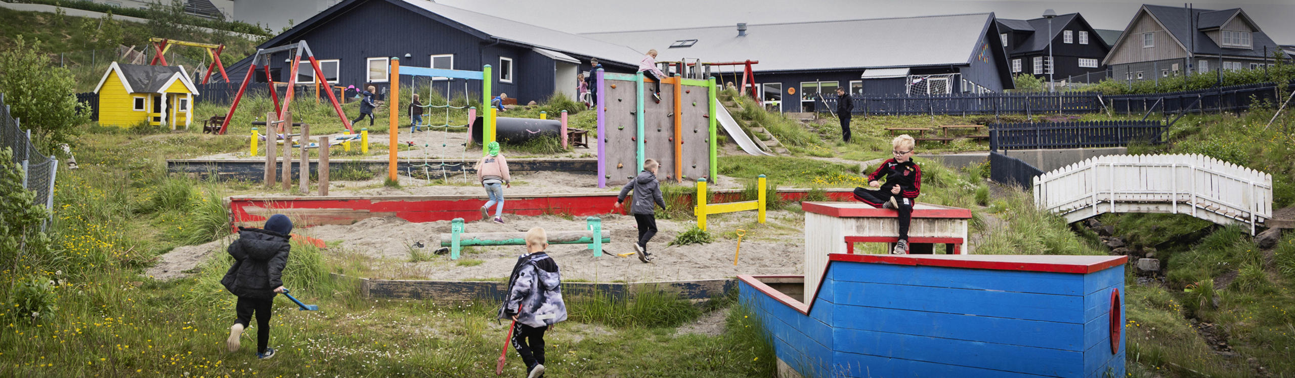 A colorful playground with children playing, featuring swings, climbing structures, and a sandbox, surrounded by greenery and buildings.