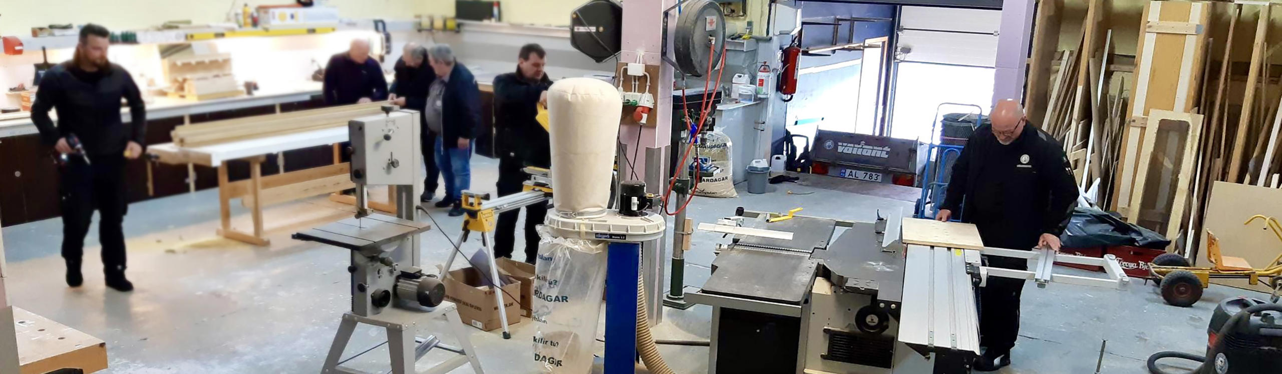 A workshop scene with several people working on woodworking projects, using various tools and machinery.