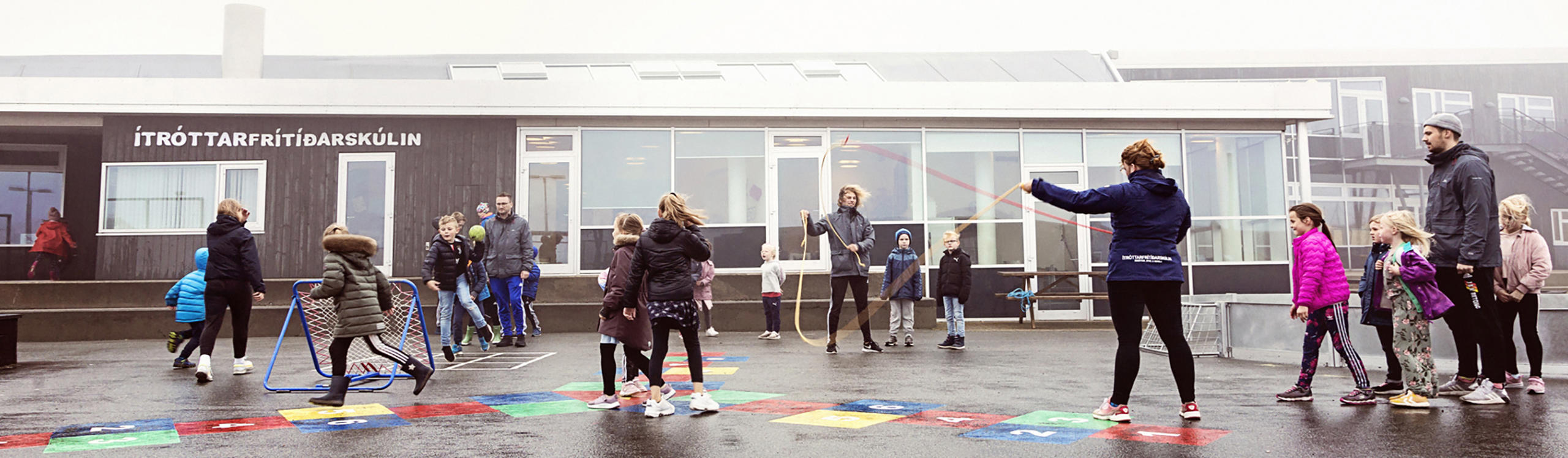 Children and adults engage in various activities outside a school building, with colorful play areas and a misty atmosphere.
