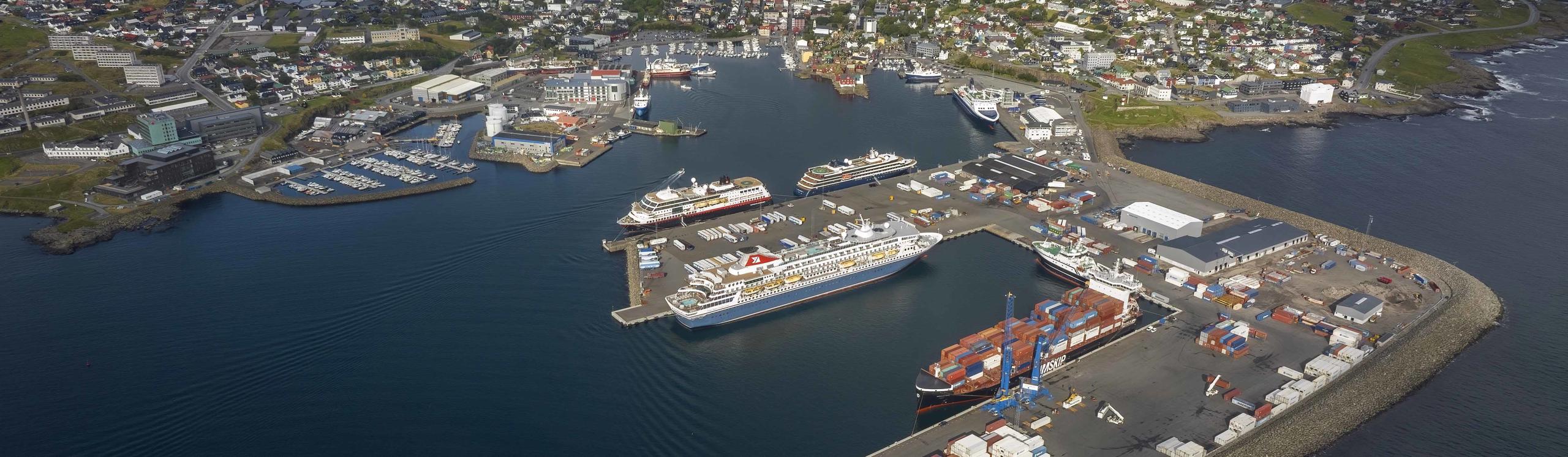 Aerial view of a harbor with several ships, docks, and surrounding buildings, set against a coastal landscape.