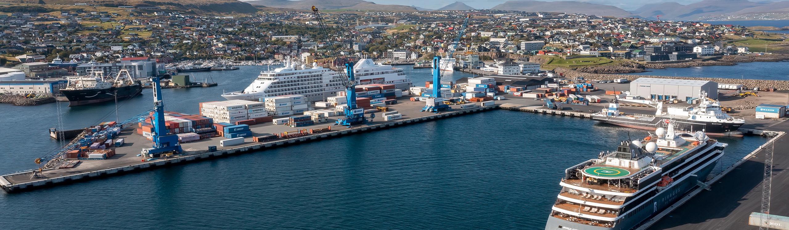 Aerial view of a busy harbor with cargo containers, ships, and a coastal town in the background.