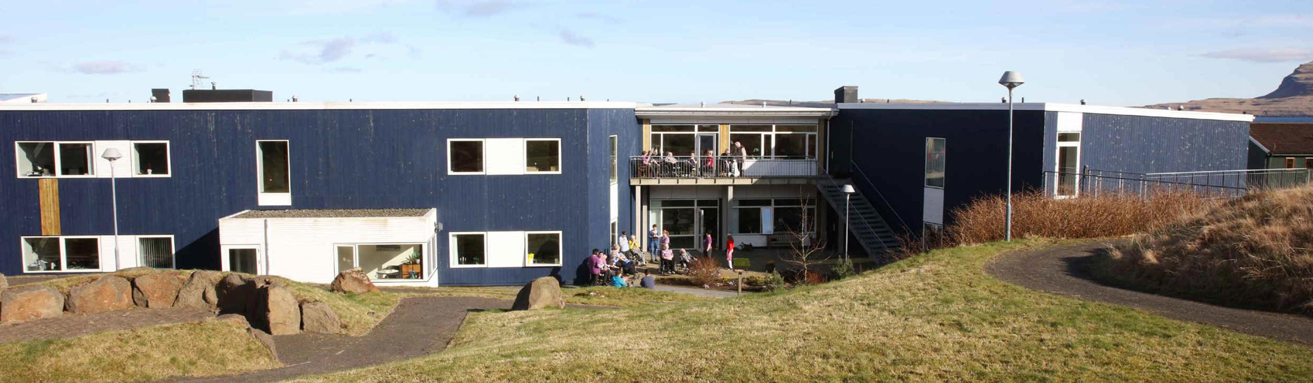 A modern blue building with multiple windows, surrounded by grass and rocks, featuring people on a balcony and in the garden area.