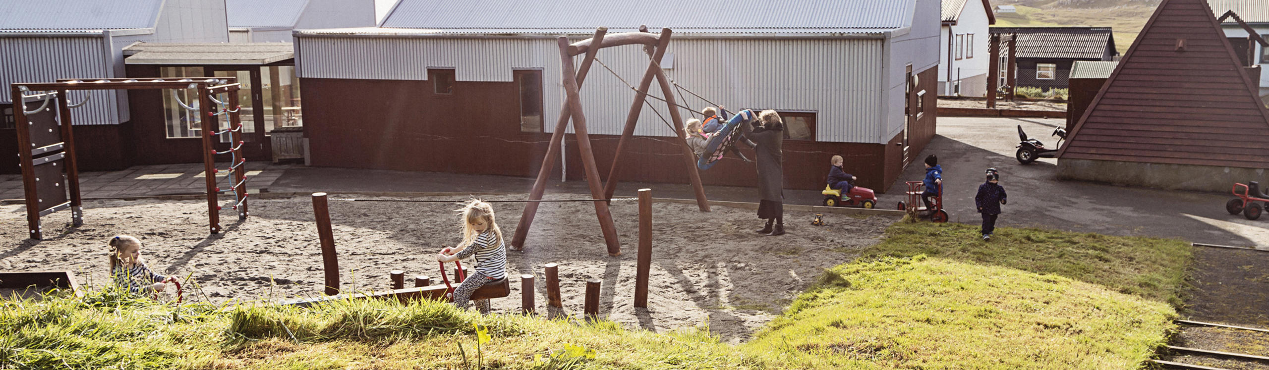 Children playing in a playground with swings and climbing structures, surrounded by buildings and grassy areas.