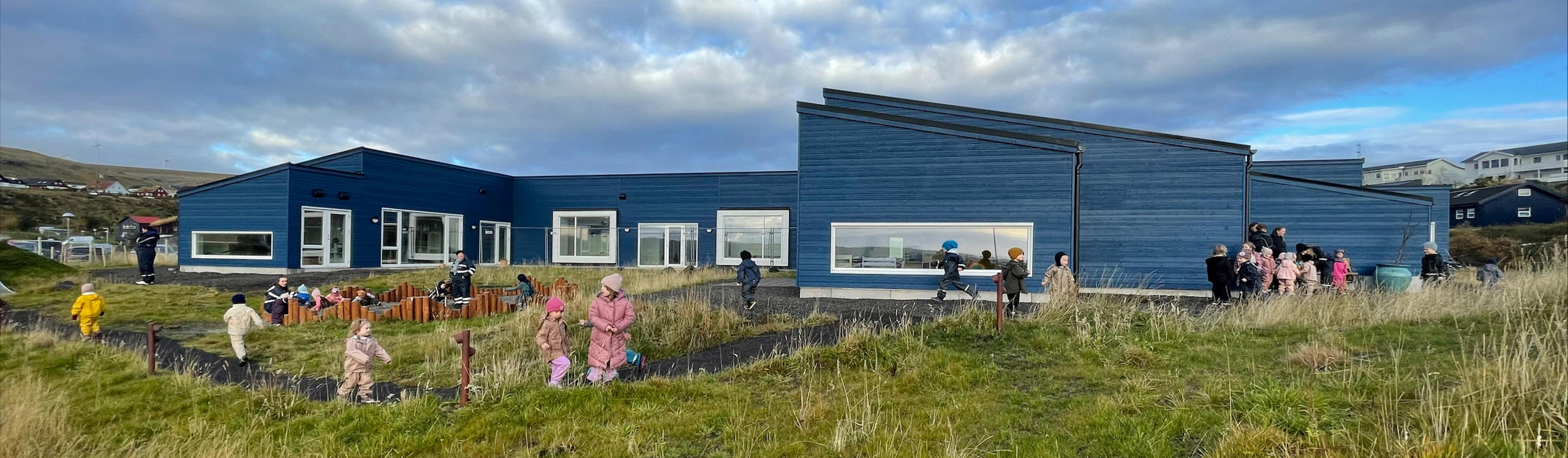 A modern blue building with children playing outside in a grassy area, under a partly cloudy sky.