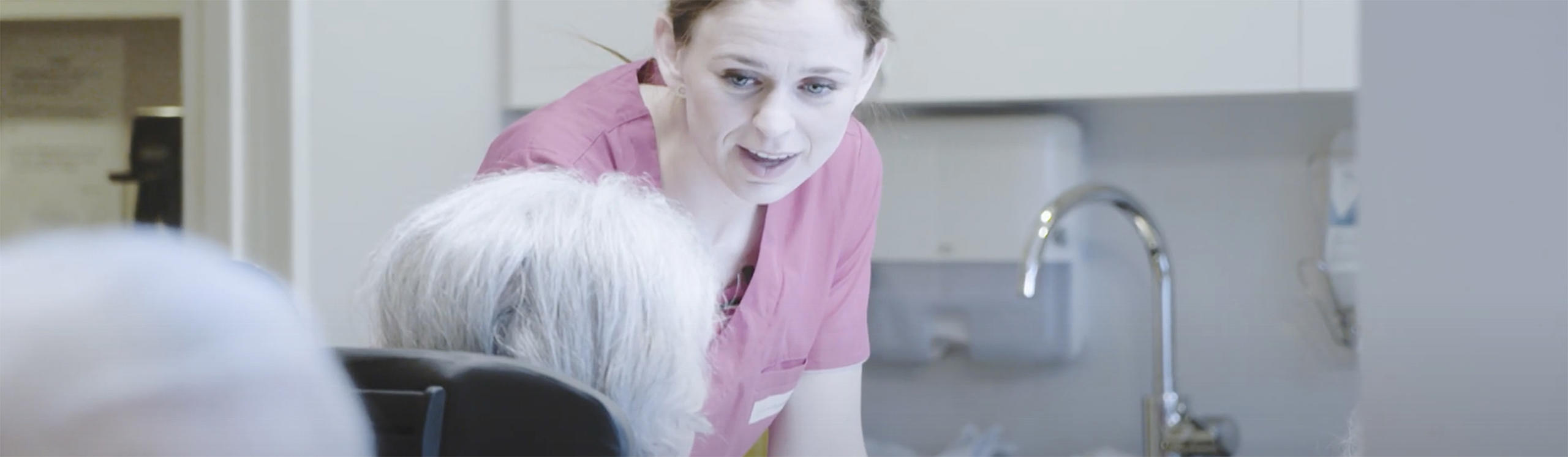 A healthcare worker in a pink uniform interacts with an elderly patient in a care setting.