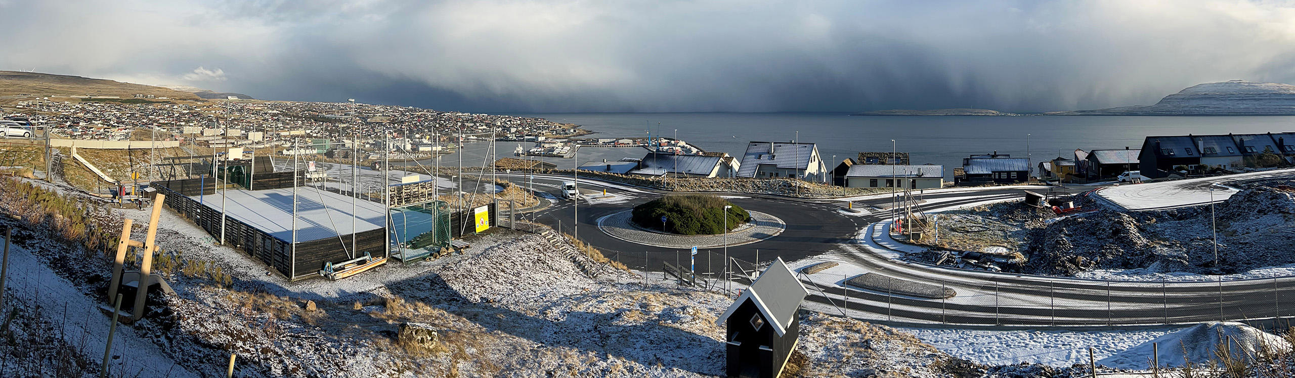 A panoramic view of a coastal town with snow-covered ground, modern buildings, and a harbor under a cloudy sky.