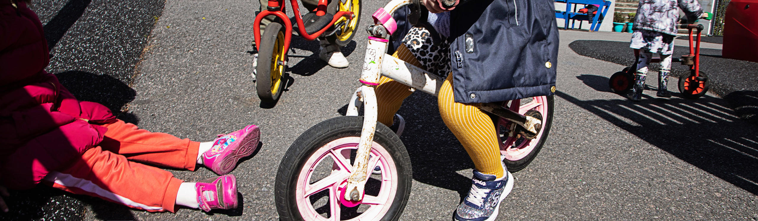 Children playing outside with tricycles and a balance bike on a sunny day. Some kids are sitting on the ground.