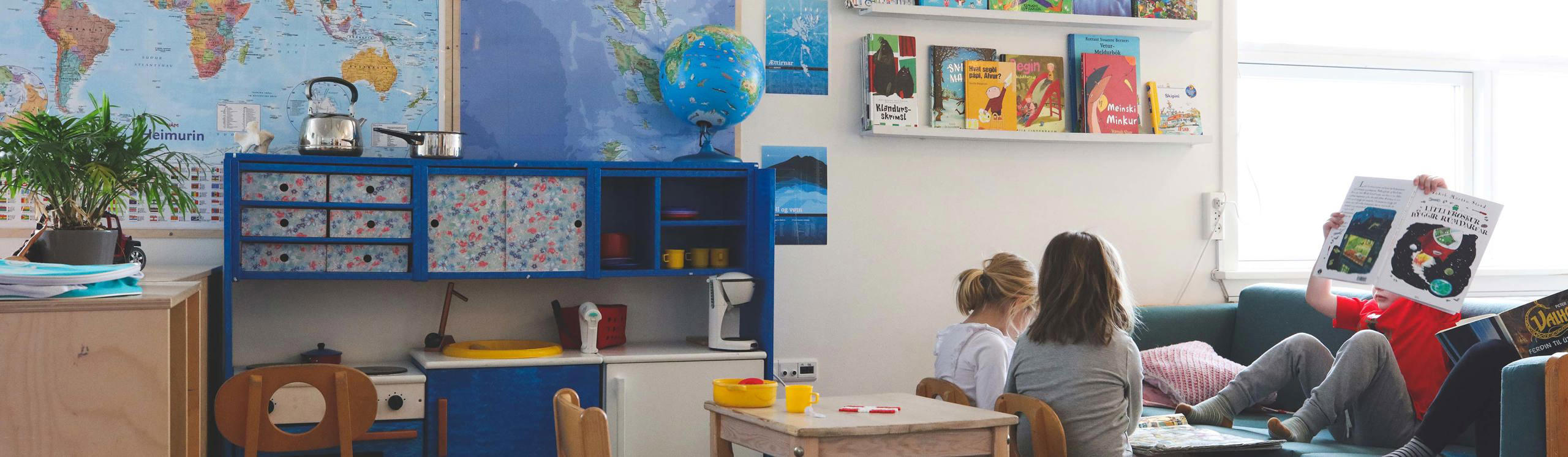A cozy room with a world map, colorful bookshelves, a play kitchen, and children reading on a couch.