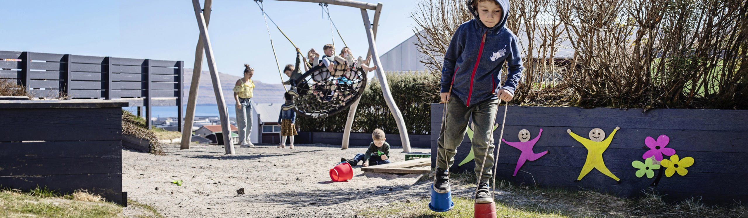 Children play in a sunny playground with swings and colorful decorations. One child balances on stilts while others enjoy the swings.
