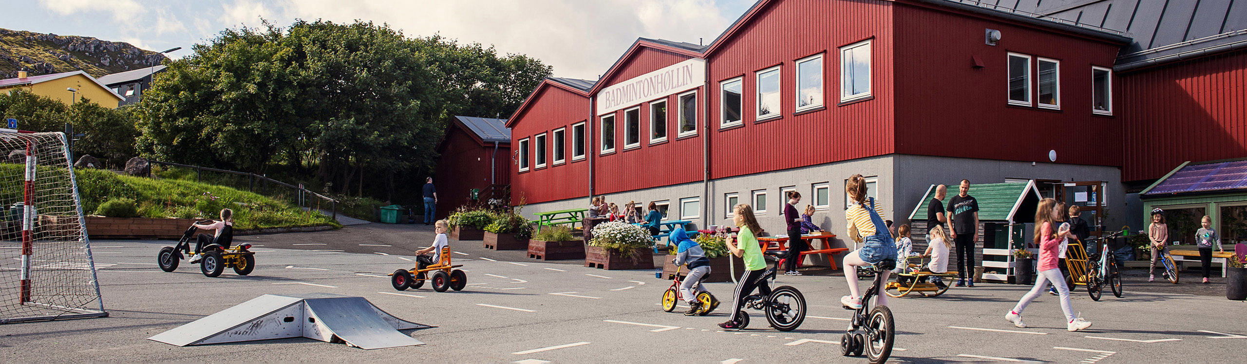 Children riding tricycles and bikes in a playground area near a red building, with a soccer goal and greenery in the background.