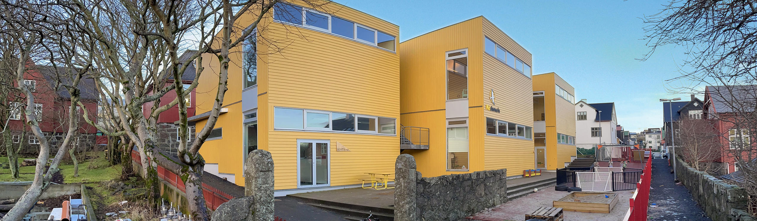 A modern yellow building surrounded by trees, with a pathway and nearby red structures in a residential area. Clear blue sky above.