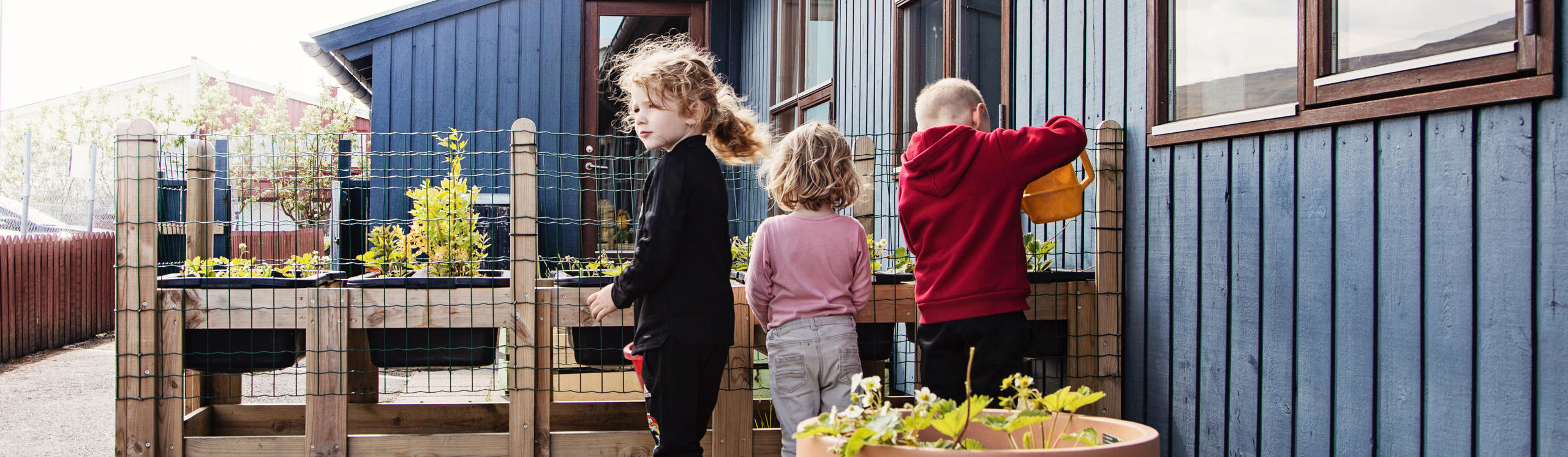 Children are gardening near a blue wooden building, tending to plants in raised beds. One child waters the plants.
