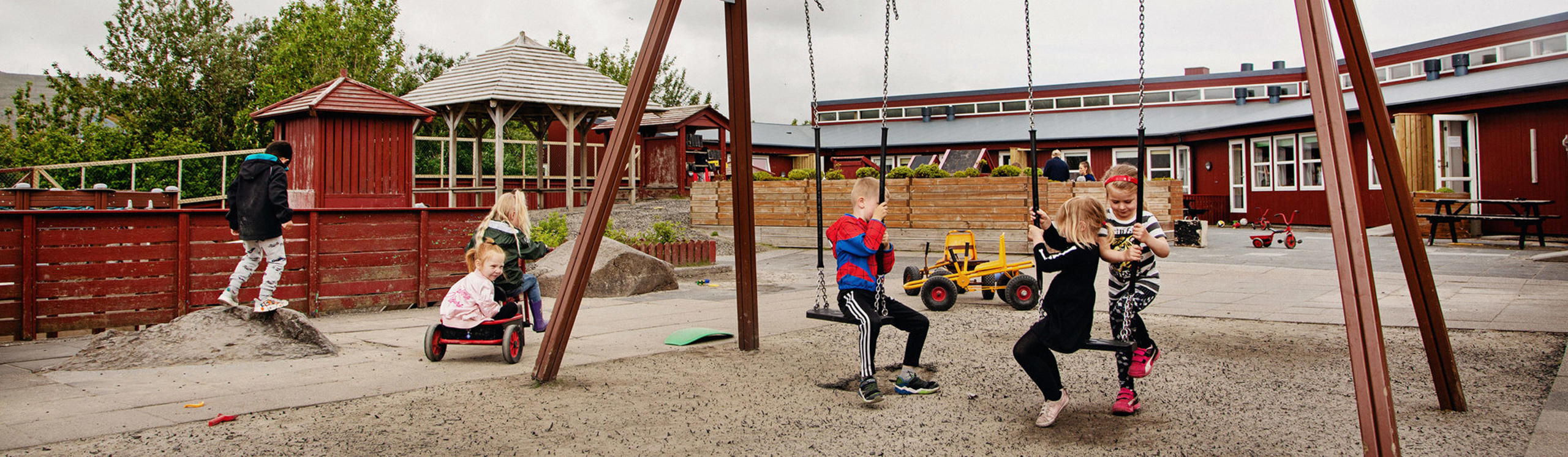 Children playing in a playground with swings, a sandbox, and a playhouse. Some are on swings, while others explore nearby.