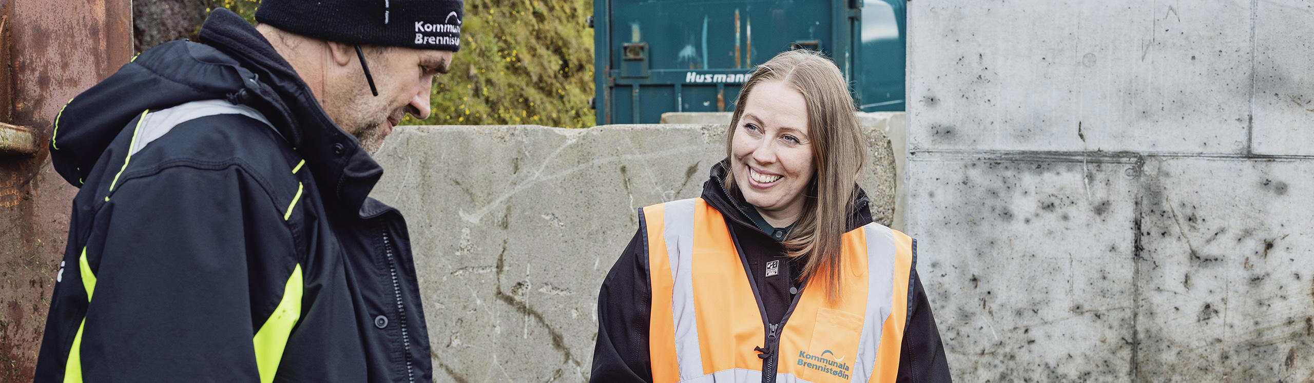 Two workers, one in an orange safety vest and the other in a black jacket, engage in conversation at a construction site.